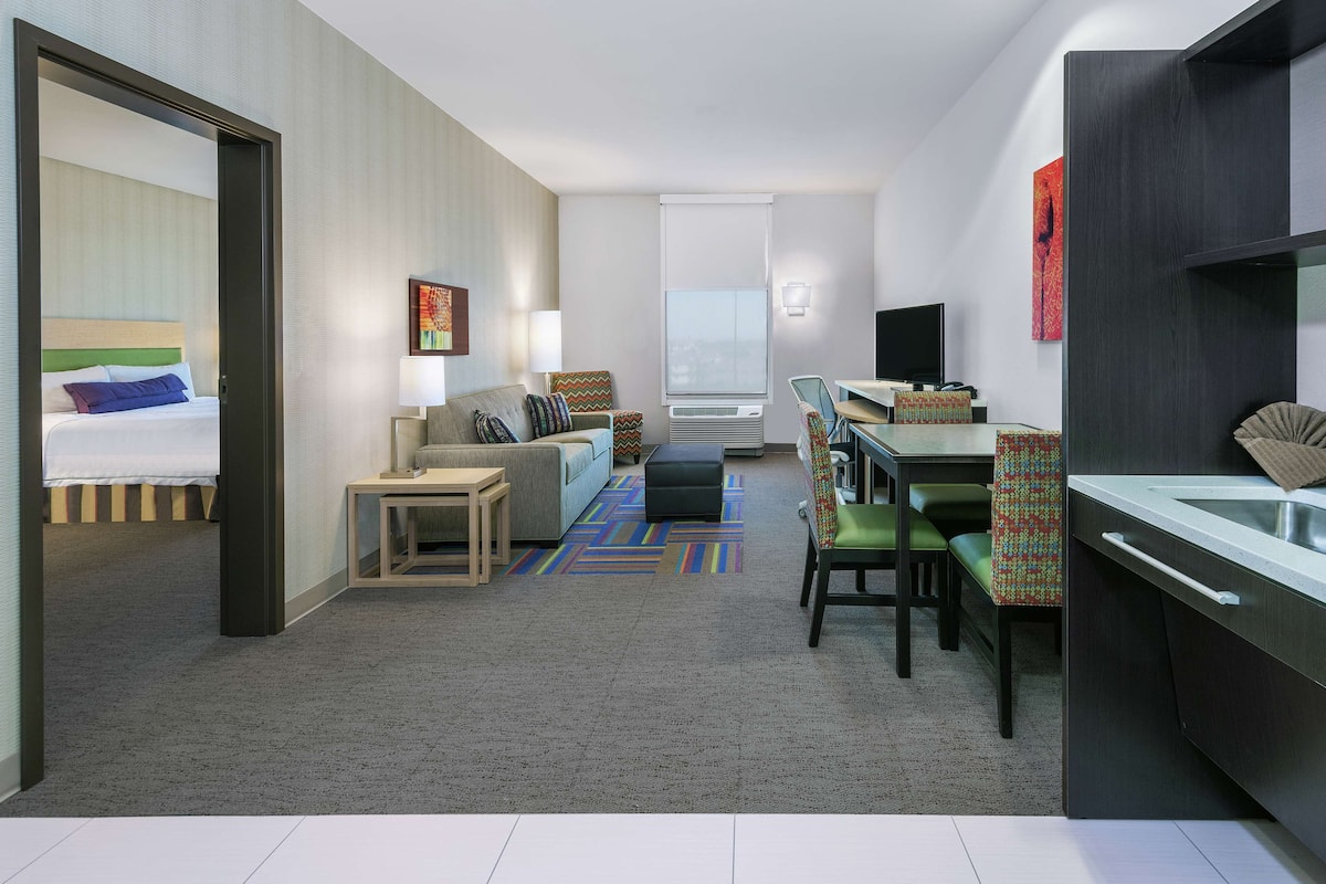A modern hotel room with a bed, a sofa, a dining area, and a kitchenette.