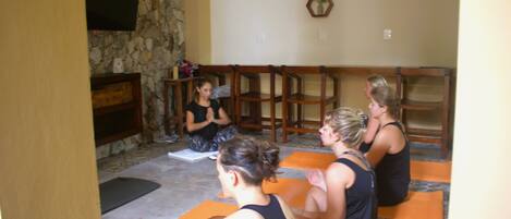 Una clase de yoga en una habitación con paredes de piedra, sillas de madera y esterillas naranjas.