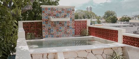 Una piscina en la azotea con una pared de azulejos y escalones de piedra, rodeada de vegetación y un paisaje urbano al fondo.