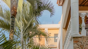 A narrow pathway flanked by large plants and white columns, leading to a building with a balcony and a staircase. - Hotel Tunich Naj (Valladolid)