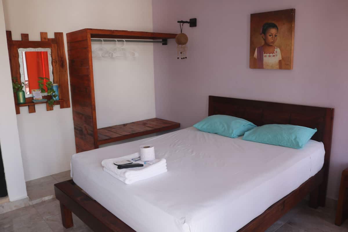 Une chambre avec un lit en bois, deux coussins turquoise, une table de chevet en bois, une étagère murale ornée de plantes, un tableau et un luminaire suspendu.