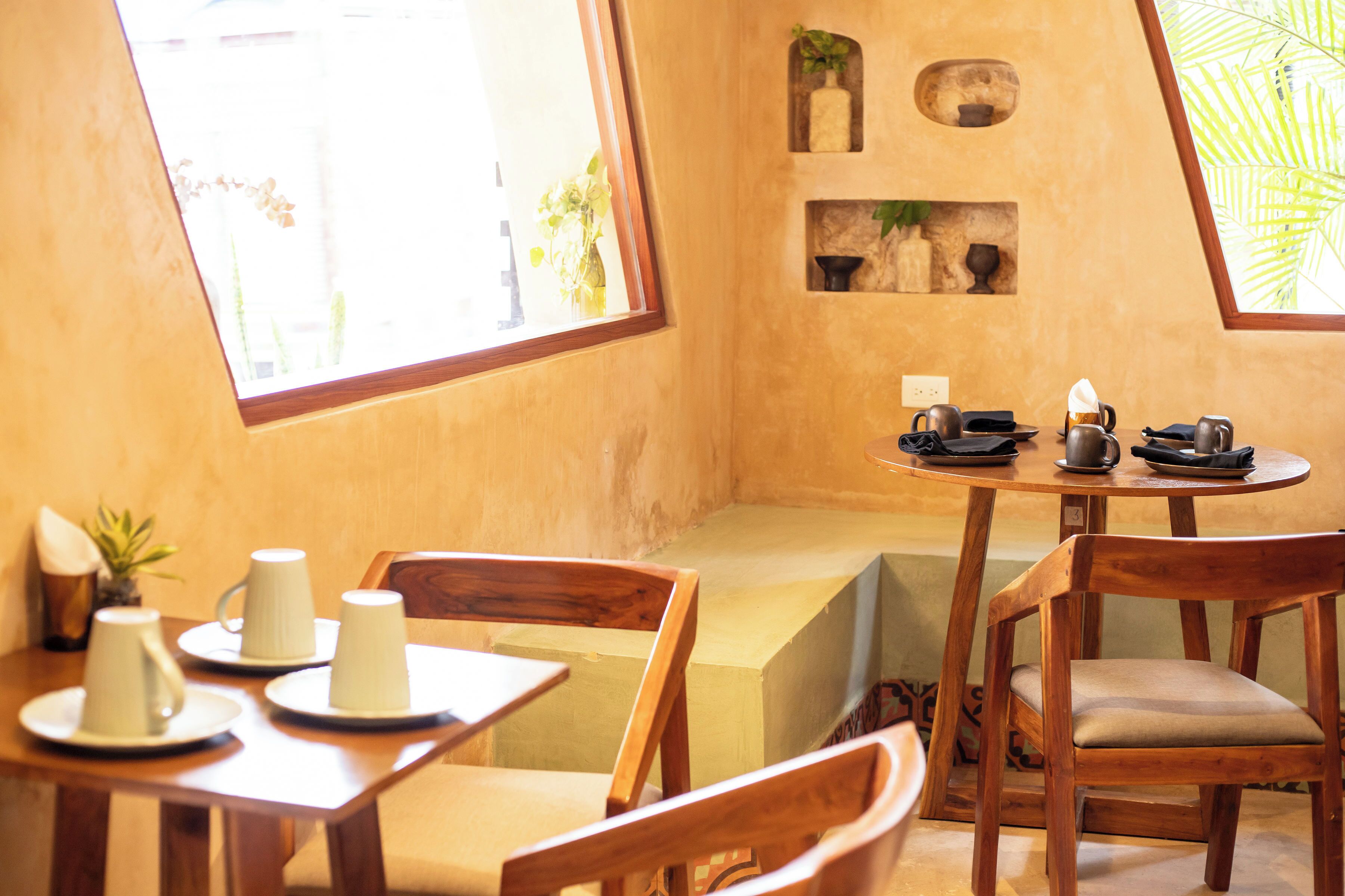 A cozy dining area with wooden furniture, a round table, and a bench. There are decorative shelves with plants and a window providing natural light.
