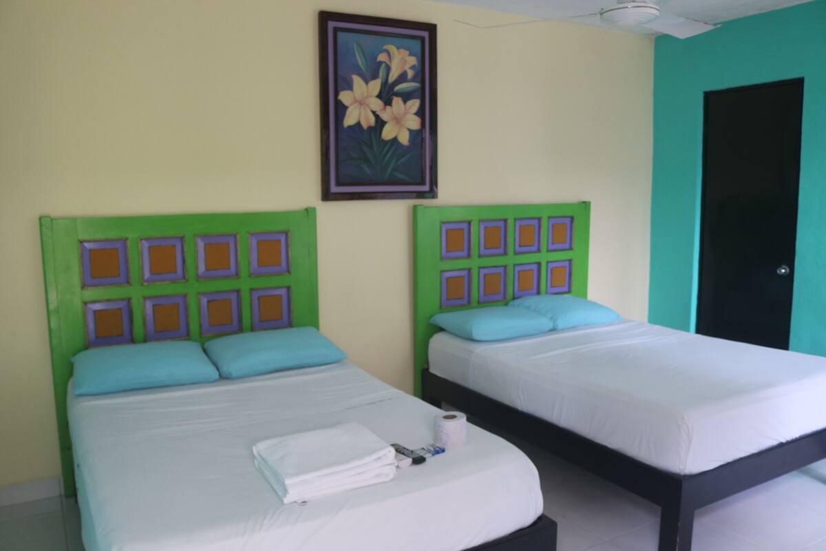 Une chambre d’hôtel avec deux lits, chacun doté d’une tête de lit verte et de draps blancs. Un tableau représentant des fleurs est accroché au mur, au-dessus des lits.