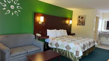 A hotel room with a green accent wall, a bed with a quilt, a sofa, a bedside table, and a nightstand with a phone.