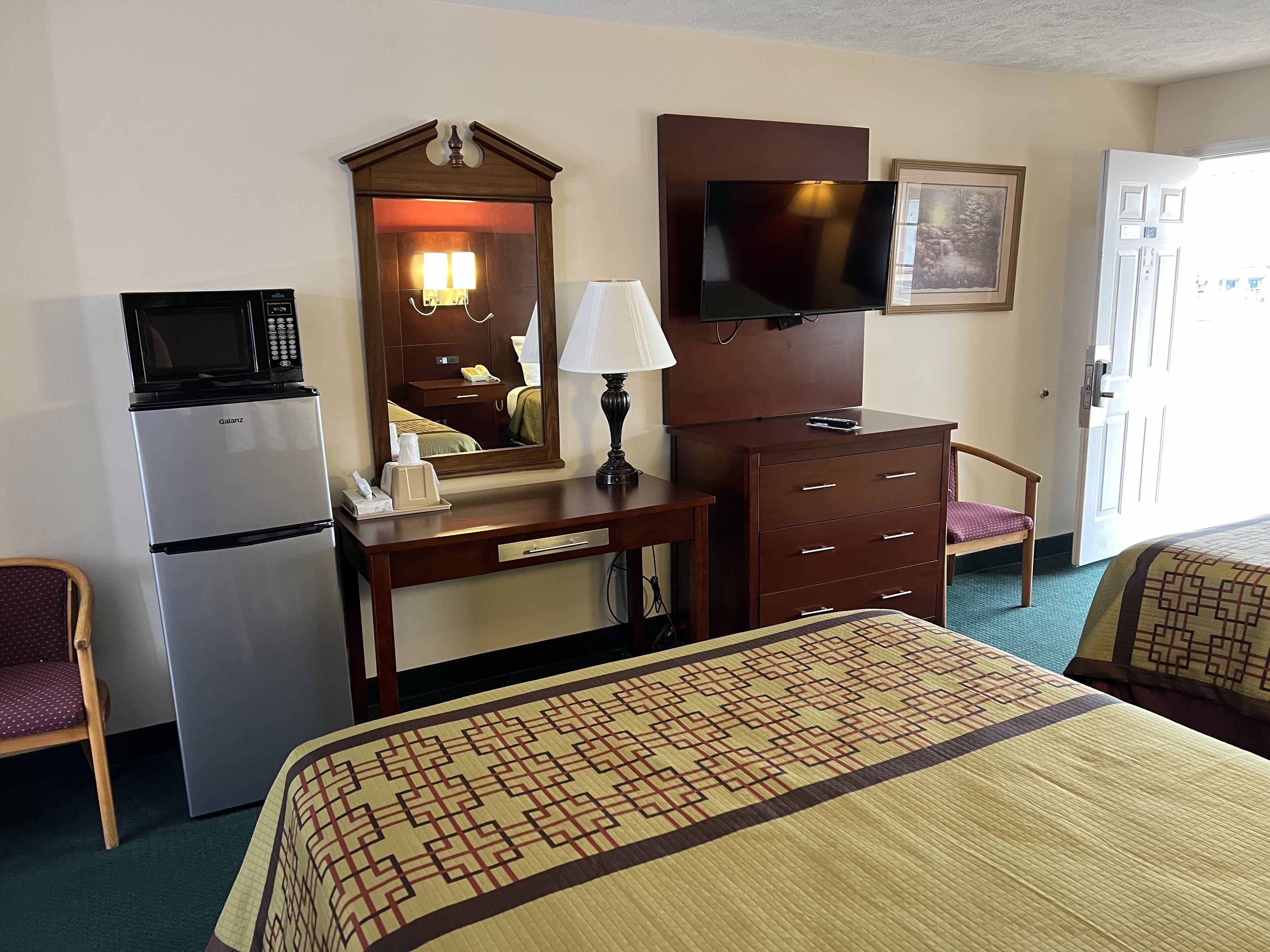 A hotel room with a bed, a desk with a lamp, a television, a microwave, and a refrigerator.