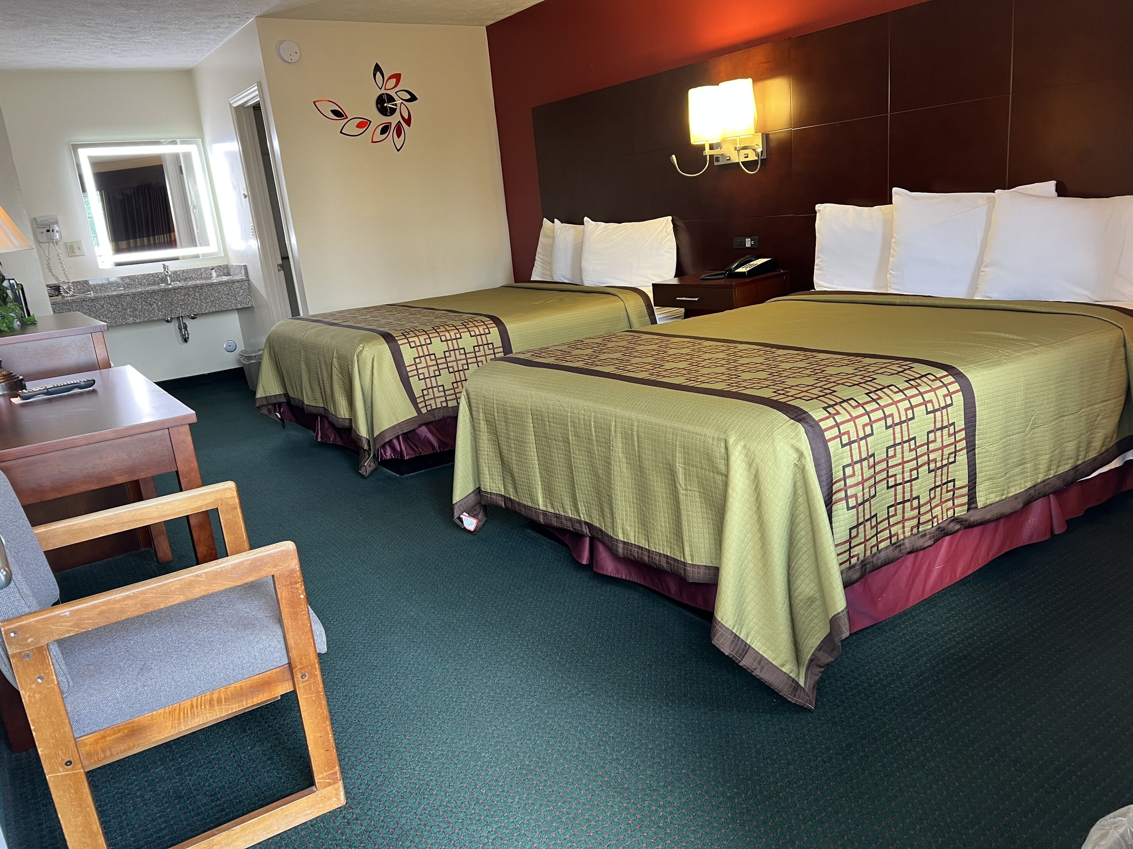 A hotel room with two beds, a desk, a chair, a small table, a mirror, a wall clock, and a nightstand with a phone.