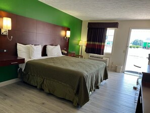 A hotel room with a large bed, a wooden headboard, a desk with a chair, a window with curtains, and a view of the outside.