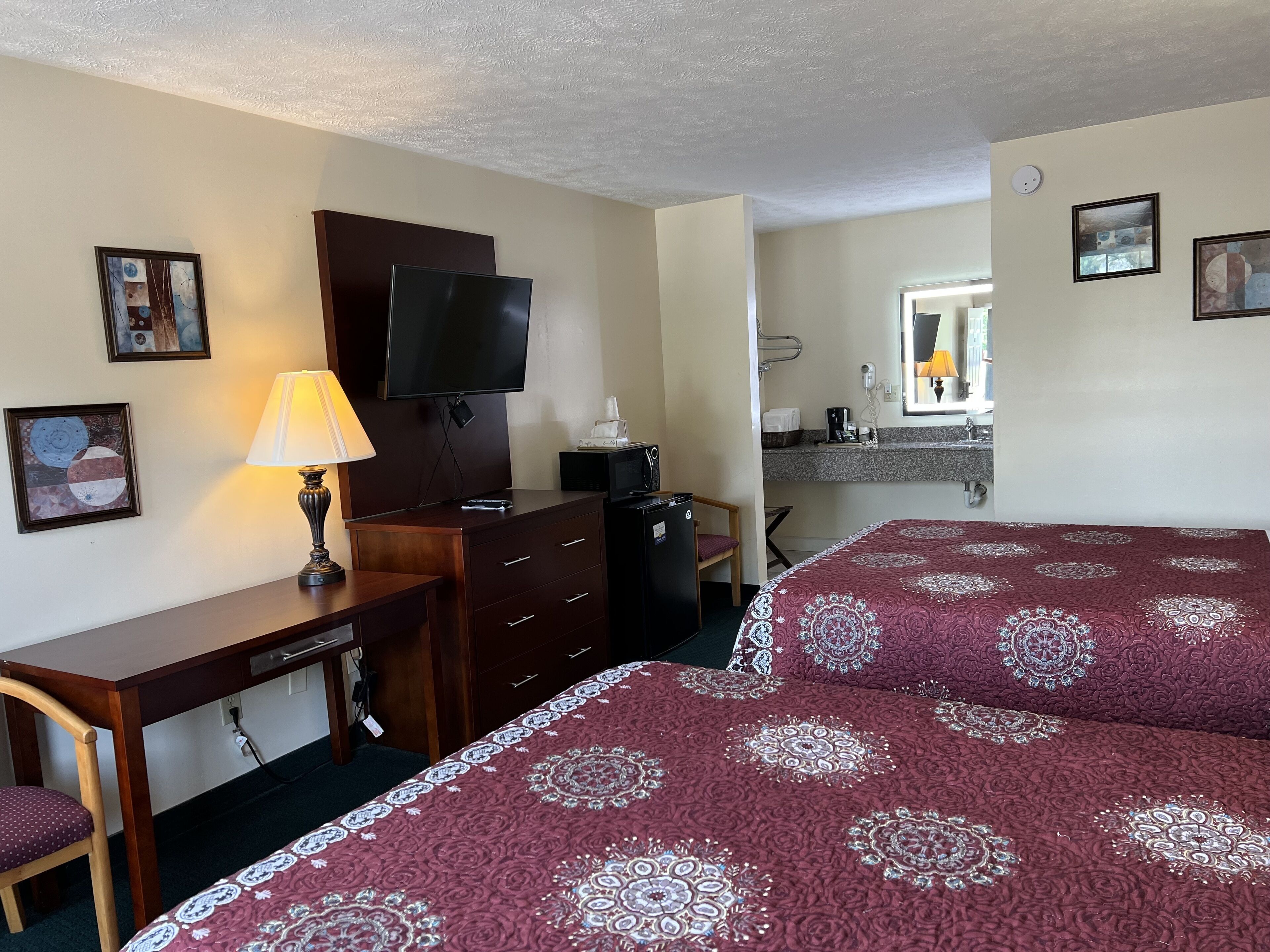 Habitación de hotel con escritorio, televisión, cama con colcha estampada, una silla y una kitchenette pequeña.