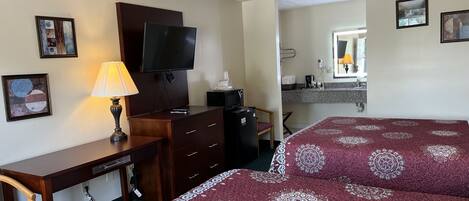 Habitación de hotel con escritorio, televisión, cama con colcha estampada, una silla y una kitchenette pequeña.