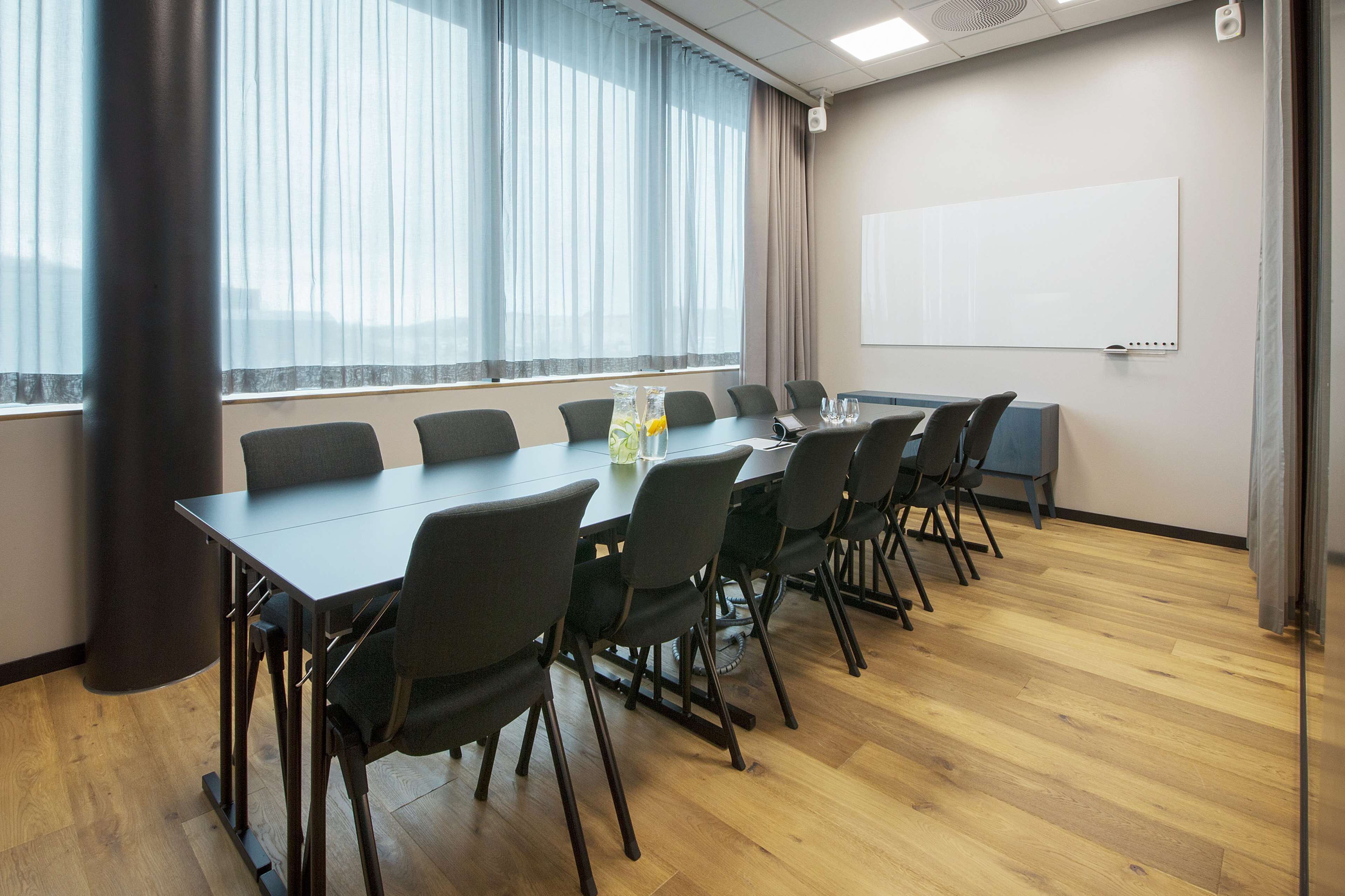 Une salle de conférence avec une longue table, des chaises, un tableau blanc et une fenêtre avec des rideaux.