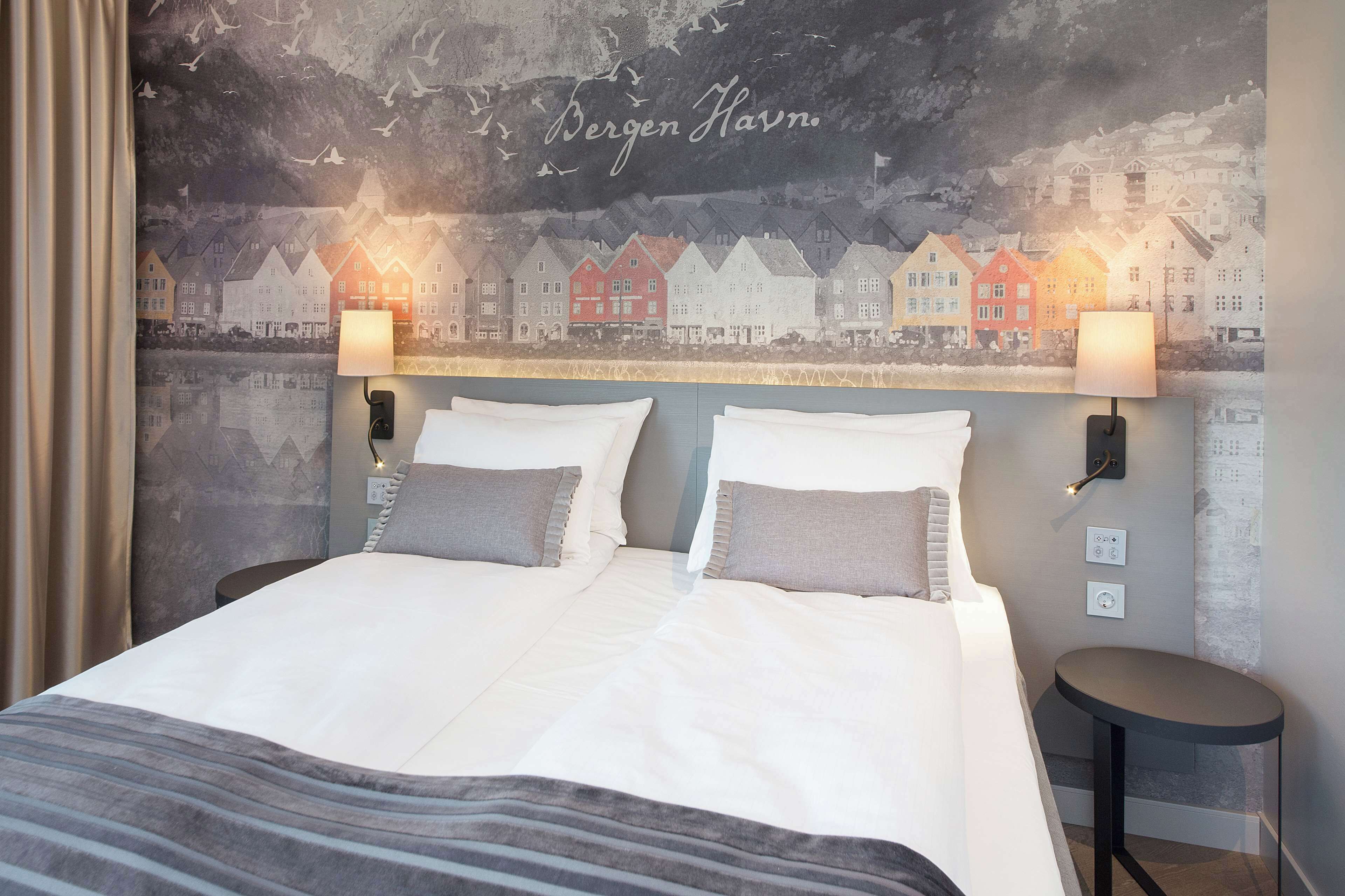 A double bed with two pillows, a bedside table, and a wall mural of houses.