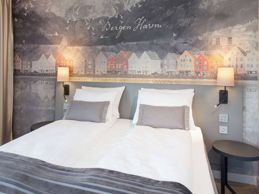 A double bed with two pillows, a bedside table, and a wall mural of houses.