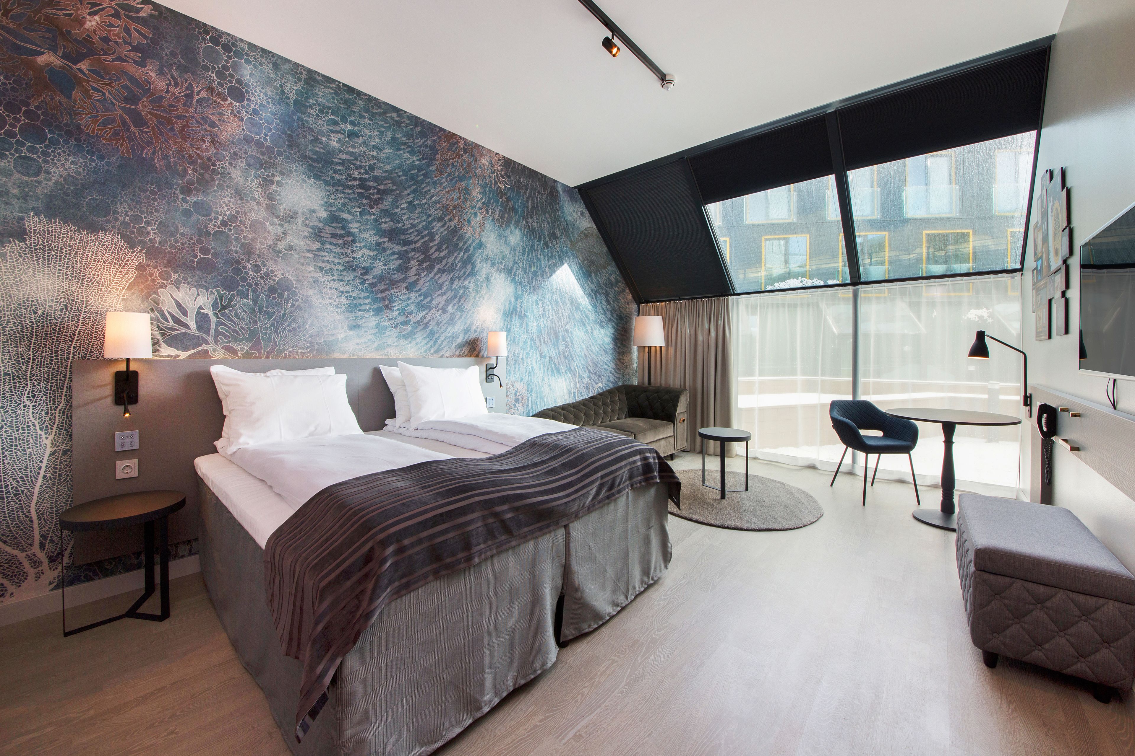 A modern hotel room with a large bed, a seating area with a round table, and a large window offering a view of the city.