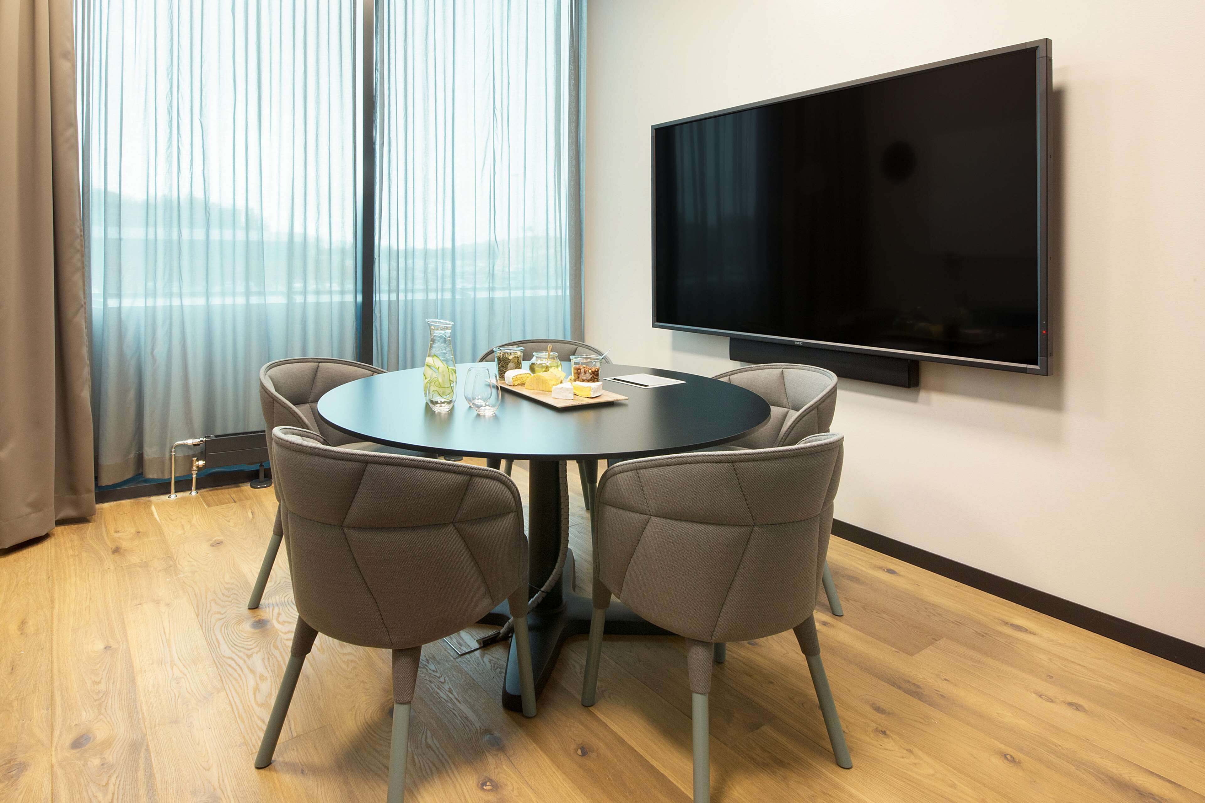 Een vergaderruimte met een ronde tafel, vier stoelen, een flatscreen televisie en een raam met gordijnen.