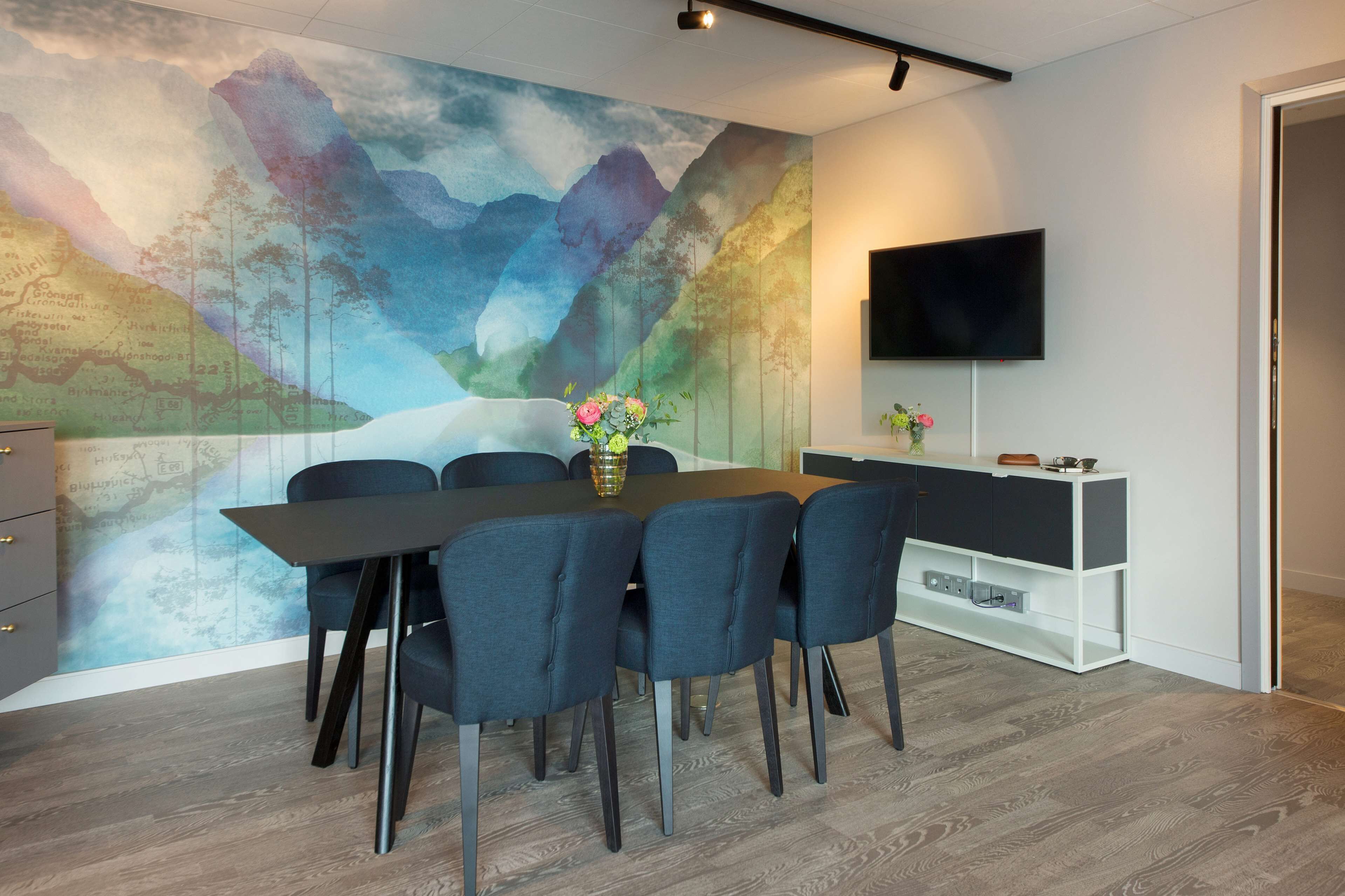 Un espace repas moderne comprenant une grande table, six chaises, un vase avec des fleurs, un téléviseur à écran plat et un meuble blanc.