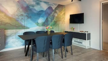 A modern dining area with a large table, six chairs, a vase with flowers, a flat-screen TV, and a white cabinet.