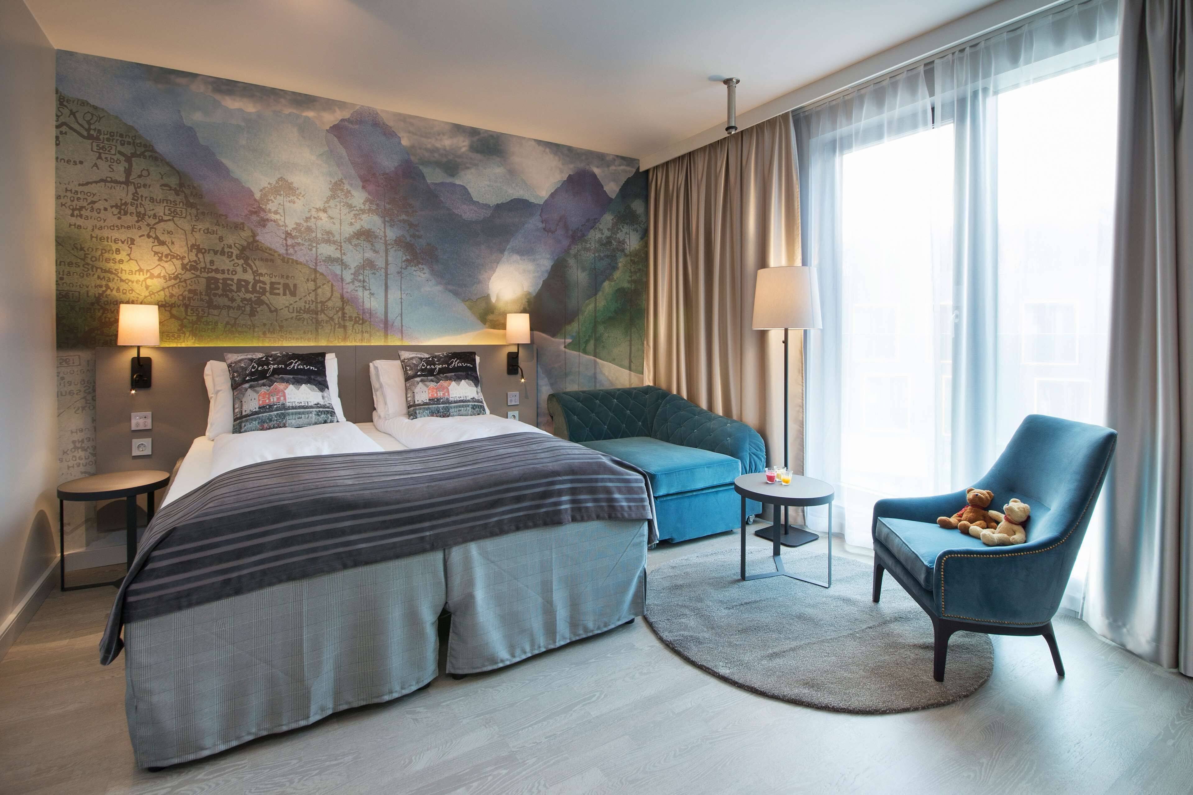 Habitación de hotel con una cama grande, un sillón azul, una mesita redonda y un mural de un mapa en la pared.