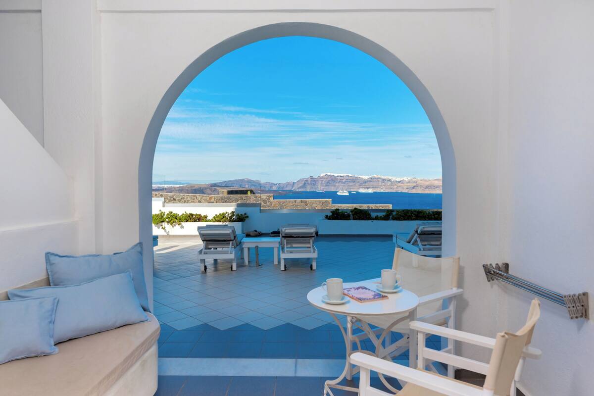 Une terrasse offrant une vue sur la mer et les montagnes, aménagée avec un canapé blanc, deux fauteuils et une petite table avec des tasses.