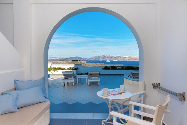 A terrace with a view of the sea and mountains, featuring a white sofa, two lounge chairs, and a small table with cups.