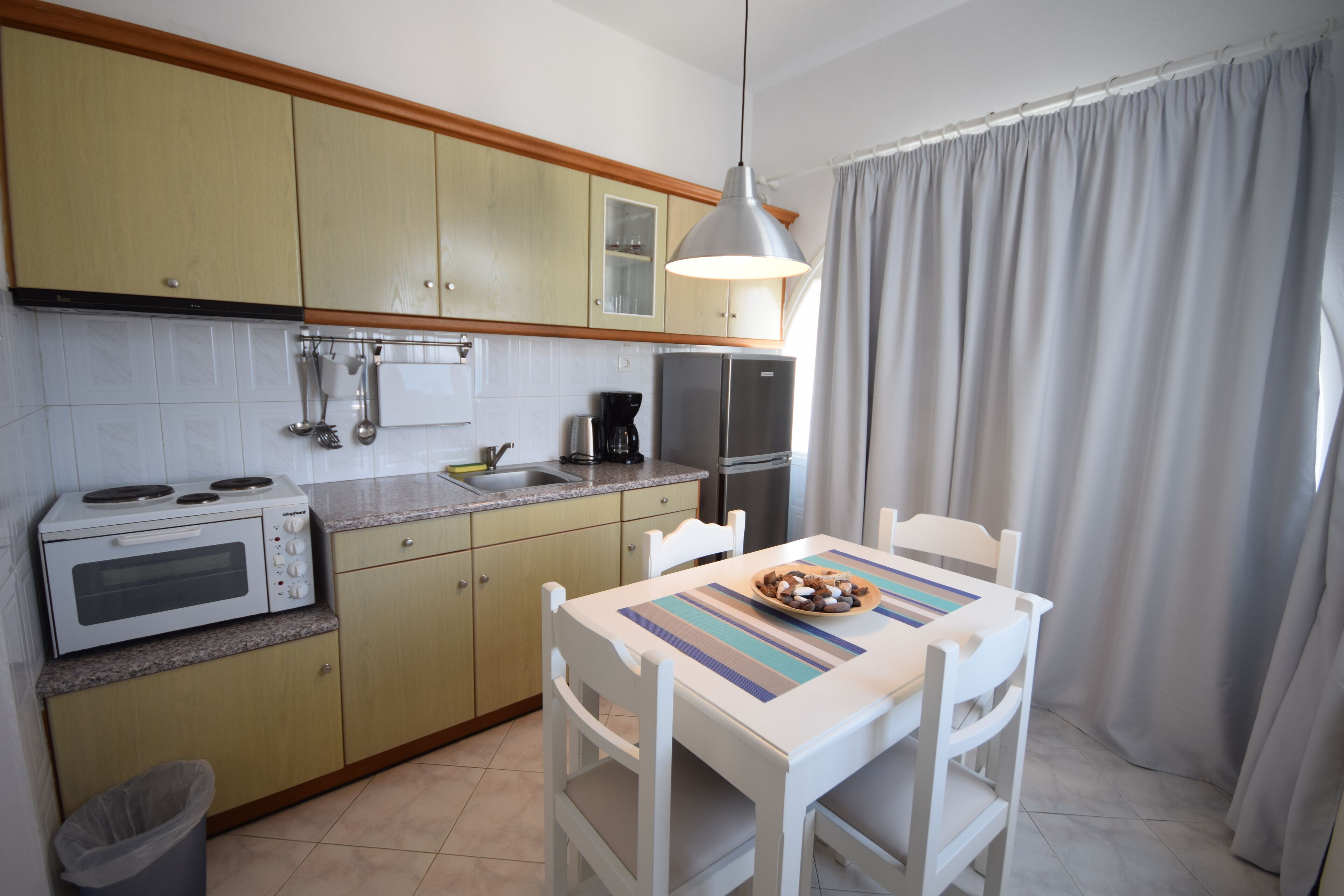 Une cuisine compacte équipée d’un four à micro-ondes, d’une cuisinière et d’un réfrigérateur. On y trouve une table avec des chaises et une fenêtre avec des rideaux.