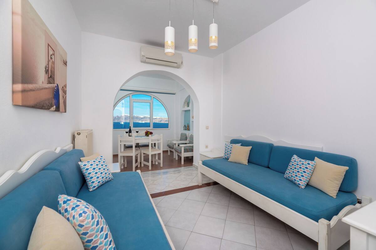 Une pièce comprenant un canapé bleu, un canapé blanc et un coin repas avec une table et des chaises. Une grande fenêtre offre une vue sur la mer.