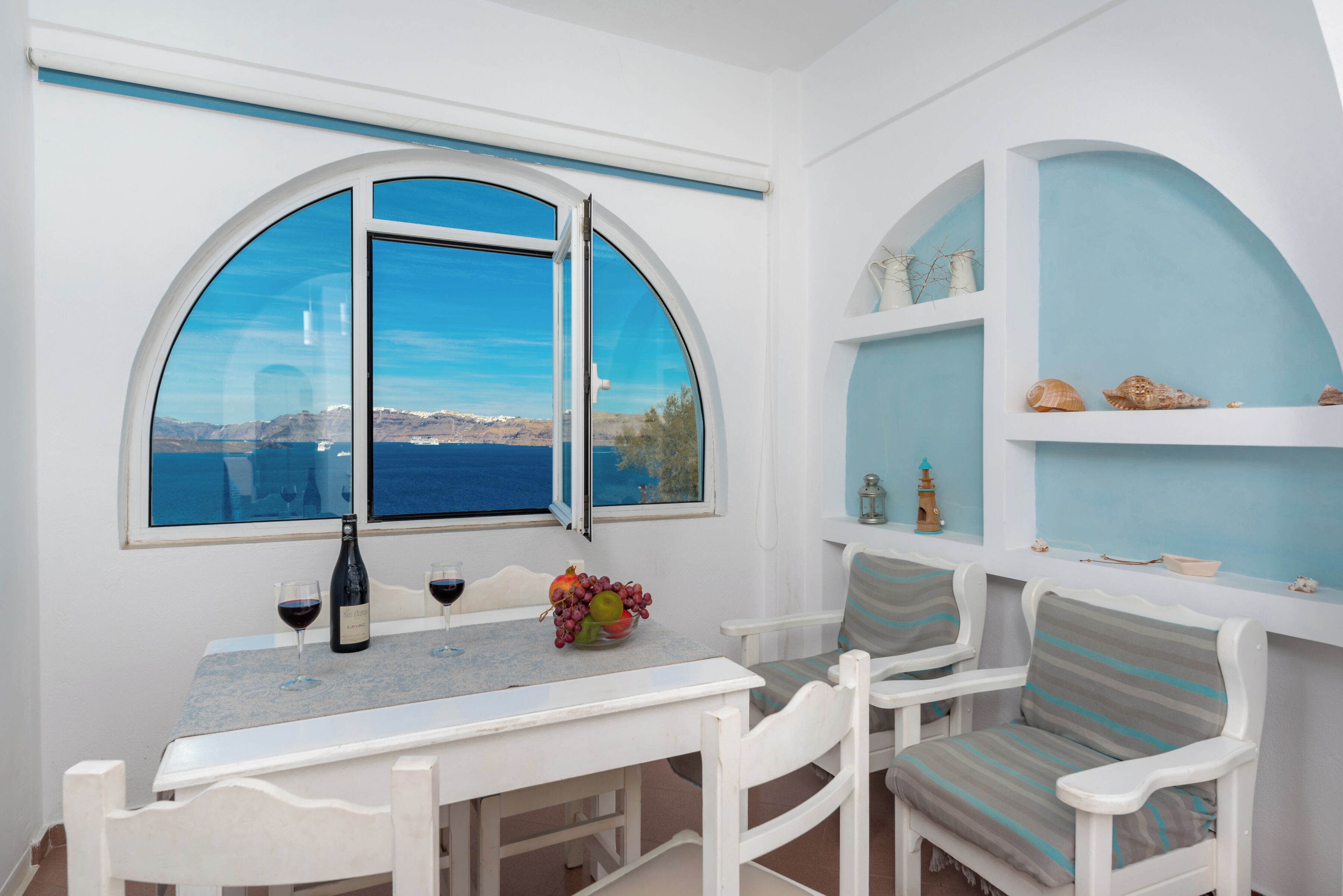 Zona pranzo con tavolo, sedie, una finestra vista mare e uno scaffale con oggetti decorativi.