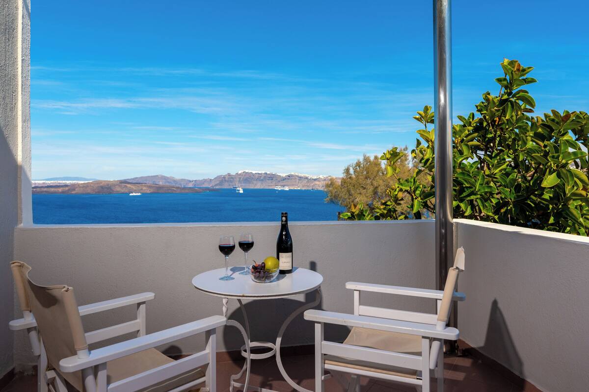 Un balcon avec des chaises blanches, une petite table ronde sur laquelle se trouvent une bouteille de vin et des verres, et une vue sur la mer et les montagnes lointaines.