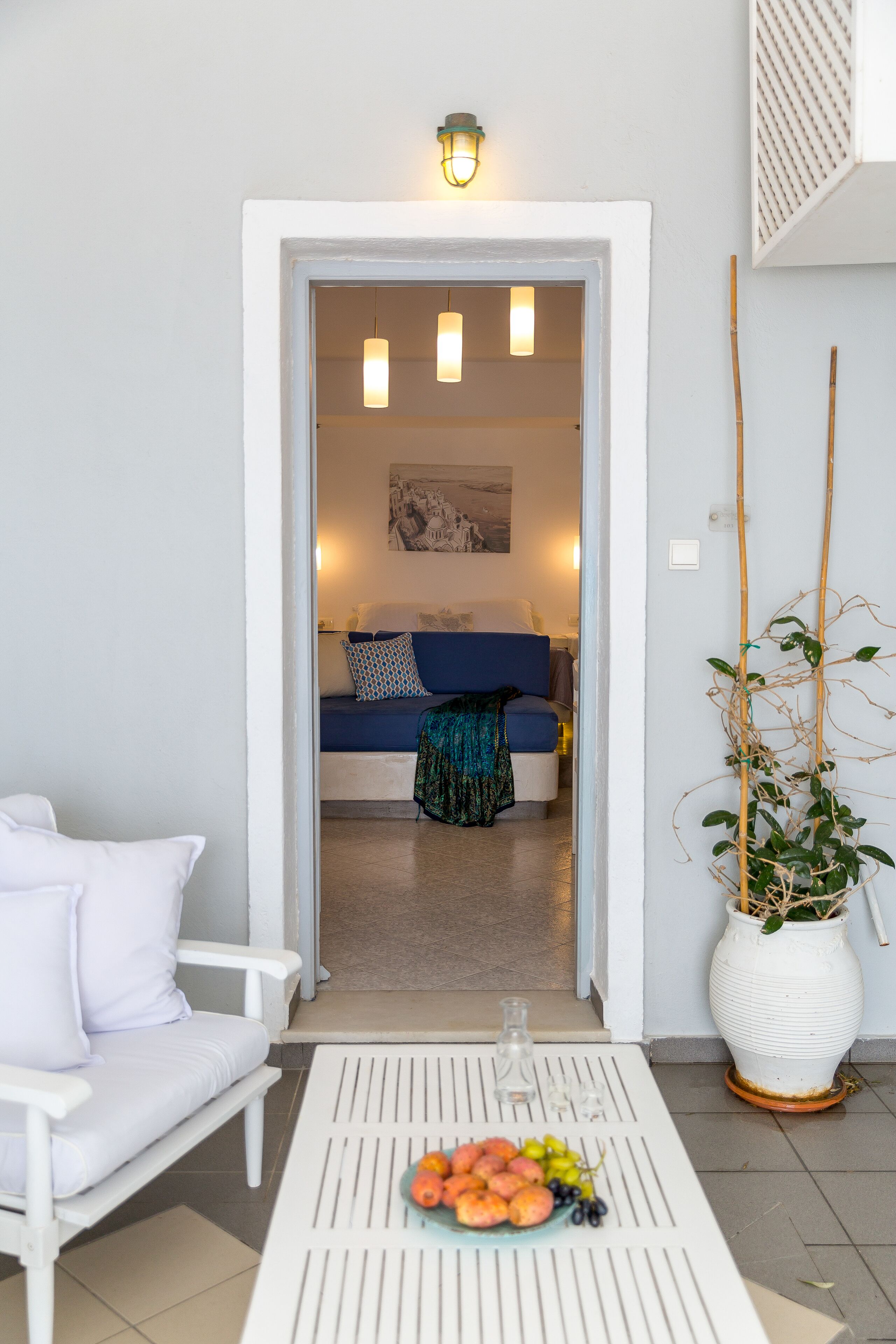 A modern room with a white sofa, a dining table with fruit, a blue sofa with patterned cushions, and a large white vase with a plant.