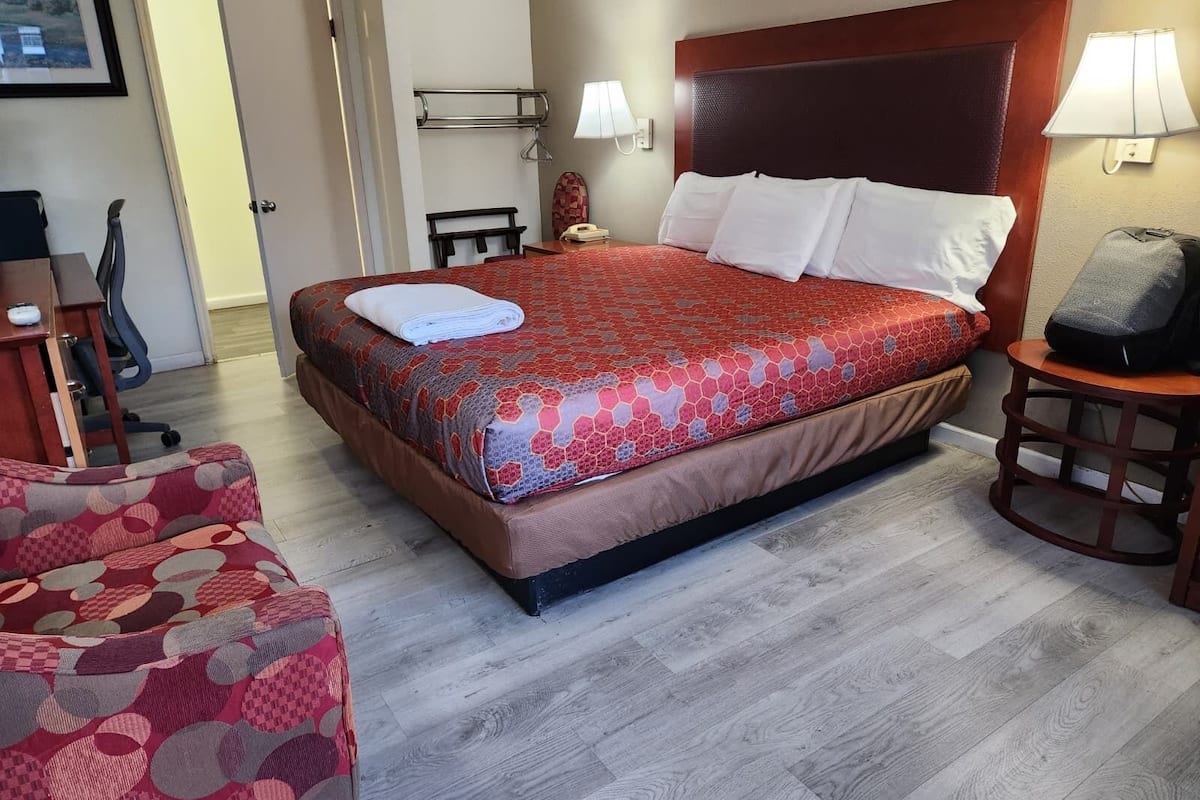Quarto de hotel com cama, uma cadeira, escrivaninha, luminária e porta.