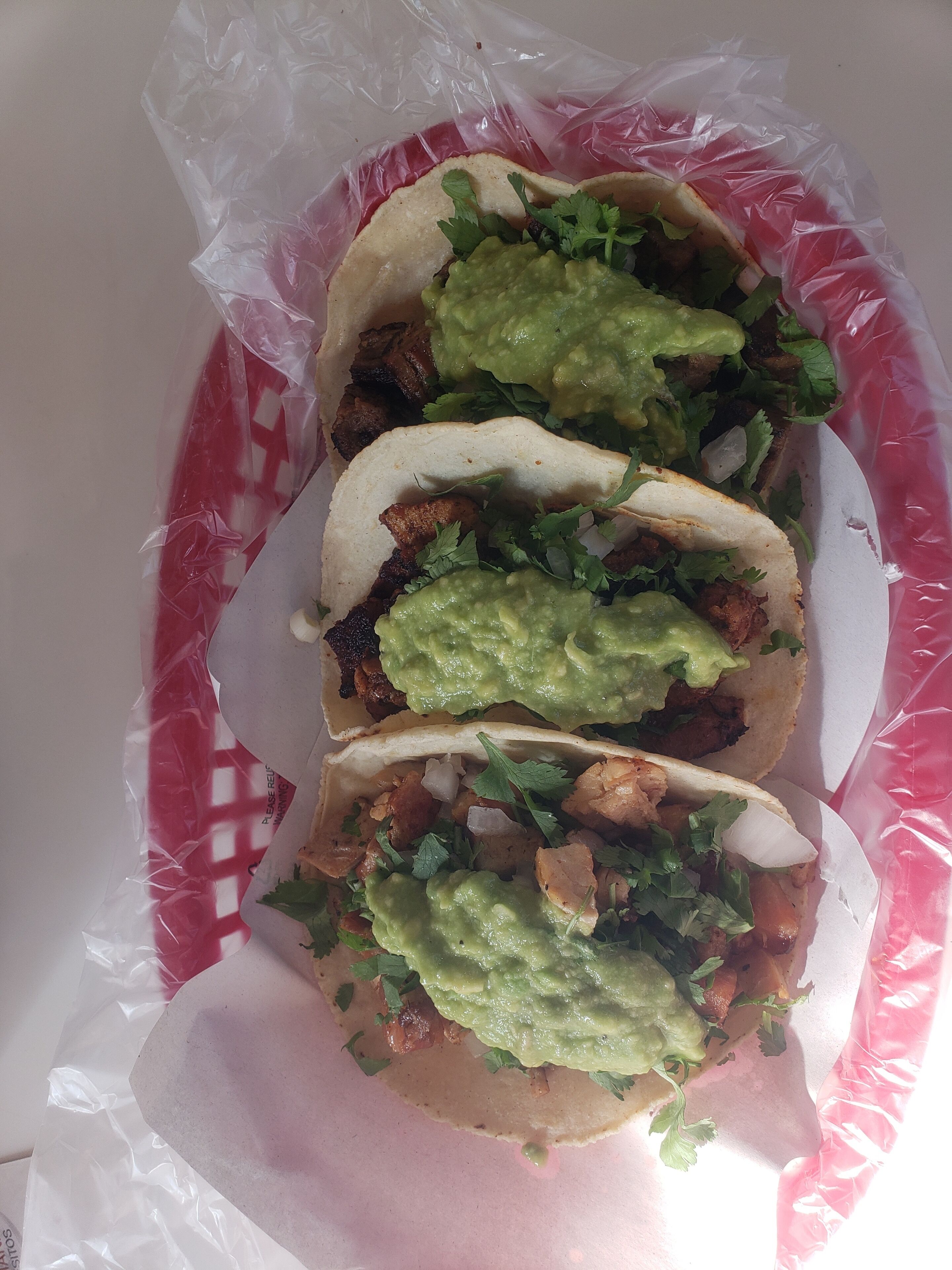 Three tacos with guacamole and cilantro, wrapped in plastic and placed on a white surface.