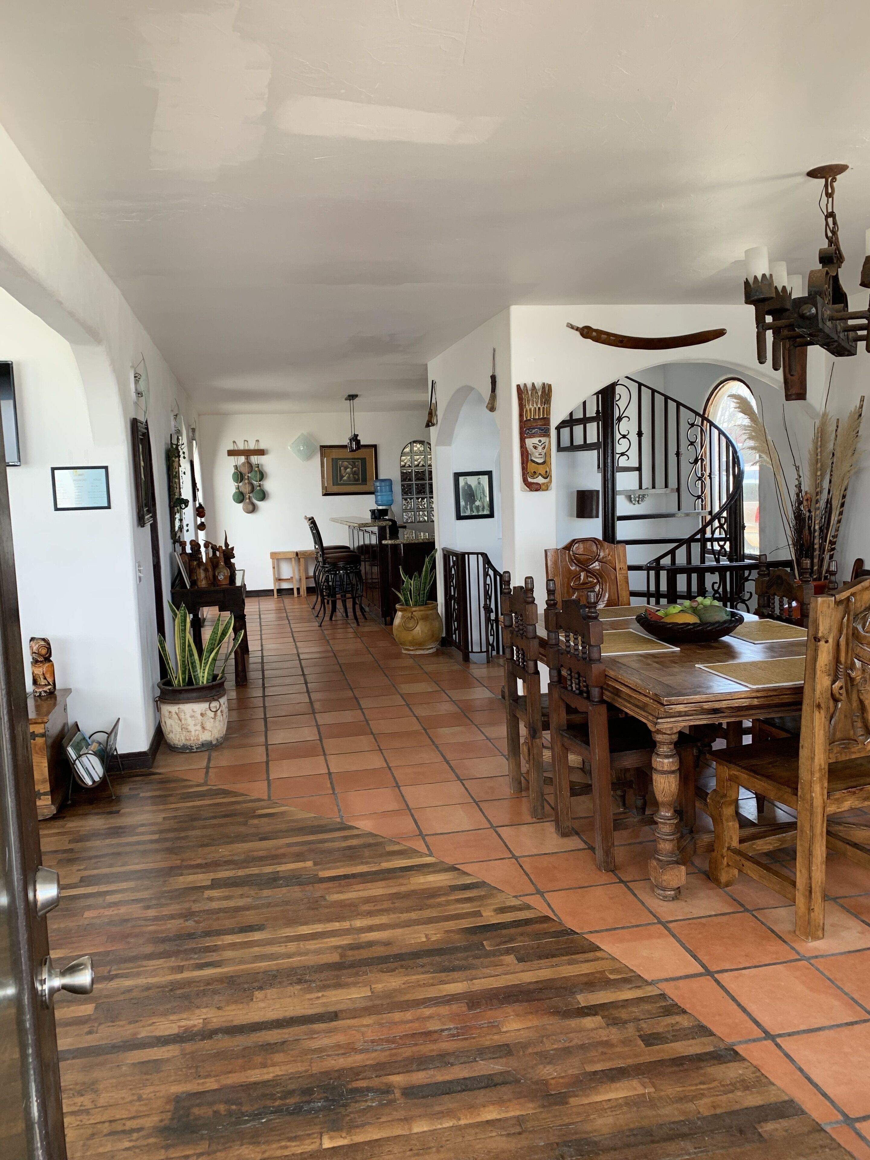 Un comedor amplio con una mesa y sillas de madera, una escalera con barandilla de hierro forjado y diversas decoraciones en las paredes, incluyendo cuadros enmarcados y una máscara grande.