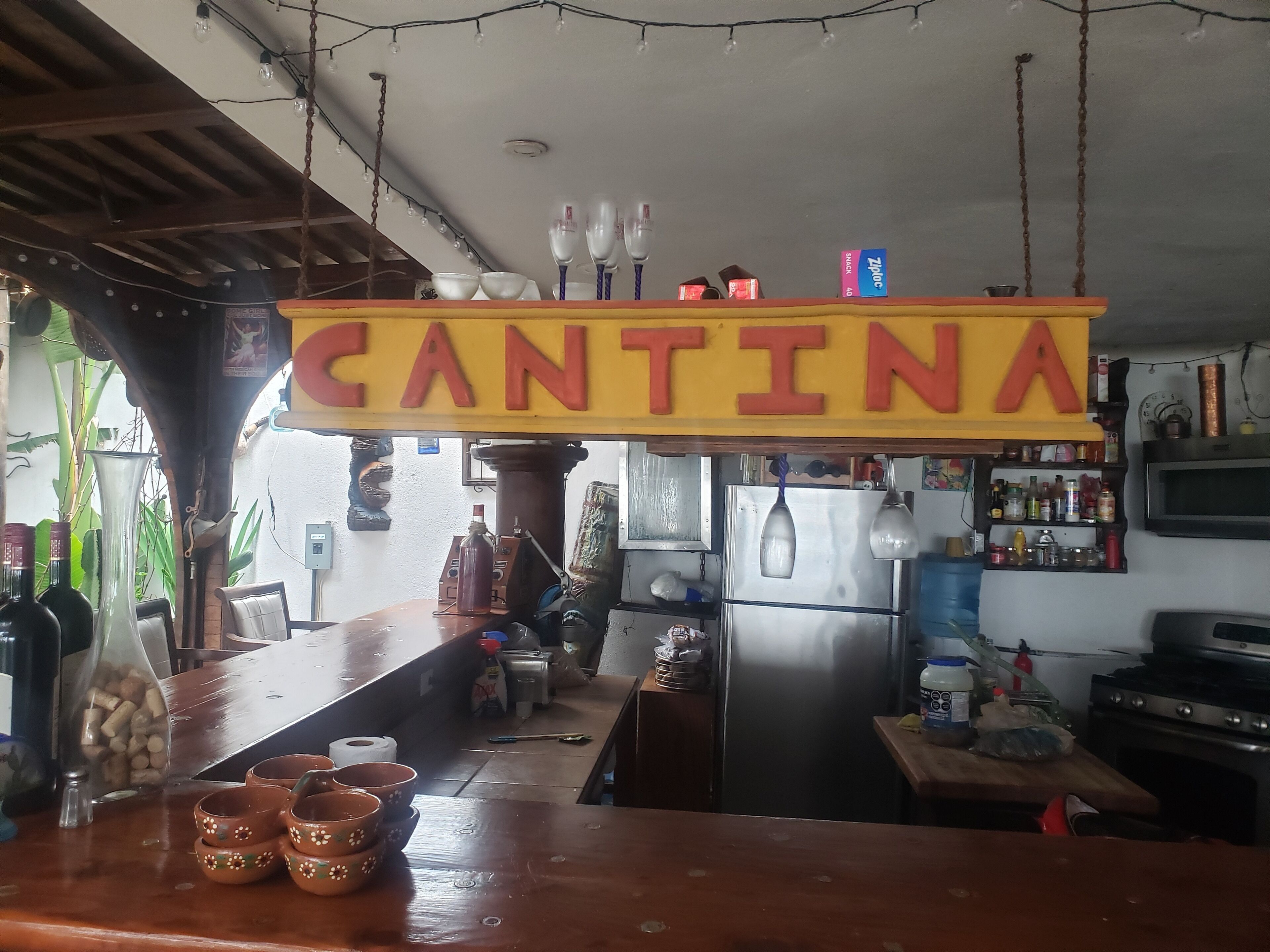 Eine Barbereich mit einem 'CANTINA'-Schild, einem Kühlschrank, Regalen voller Flaschen und einer Holztresen mit Dekoration.