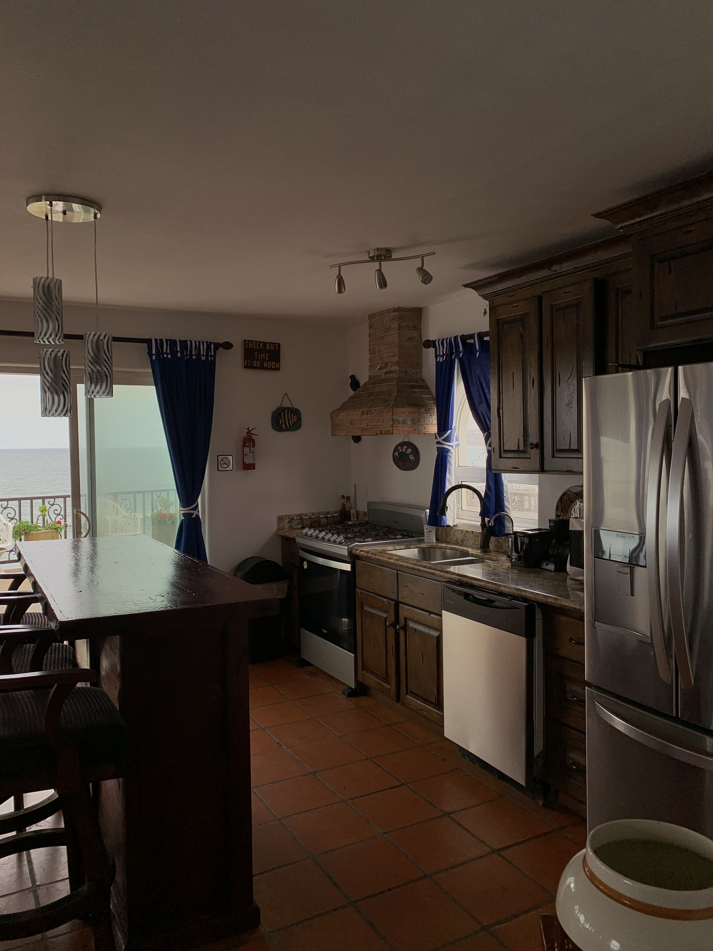 Eine Küche mit einer Kochinsel, einem Herd, Backofen, Geschirrspüler und einem Kühlschrank. Es gibt ein Fenster mit Vorhängen, einen Feuerlöscher und ein Schild an der Wand.