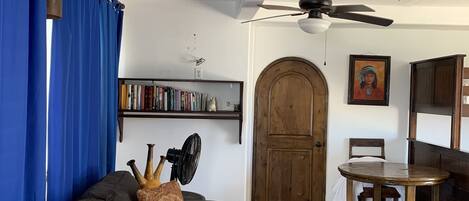 A living room with a sofa, a fan, a ceiling fan, a wooden door, a bookshelf, and a table.