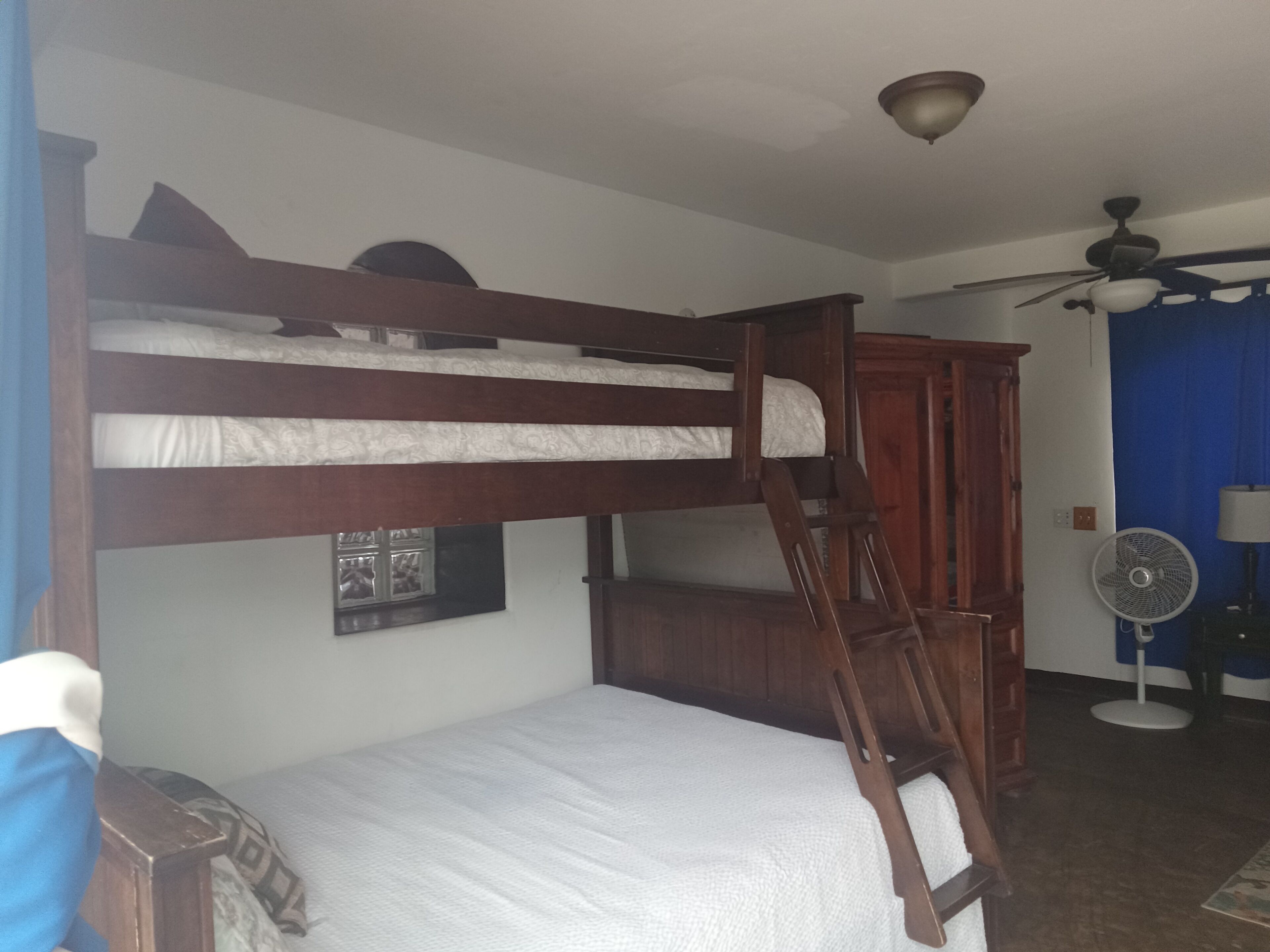 Una cama litera con escalera, un armario de madera, un ventilador de techo y una ventana con un marco de fotos.