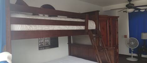 Una cama litera con escalera, un armario de madera, un ventilador de techo y una ventana con un marco de fotos.