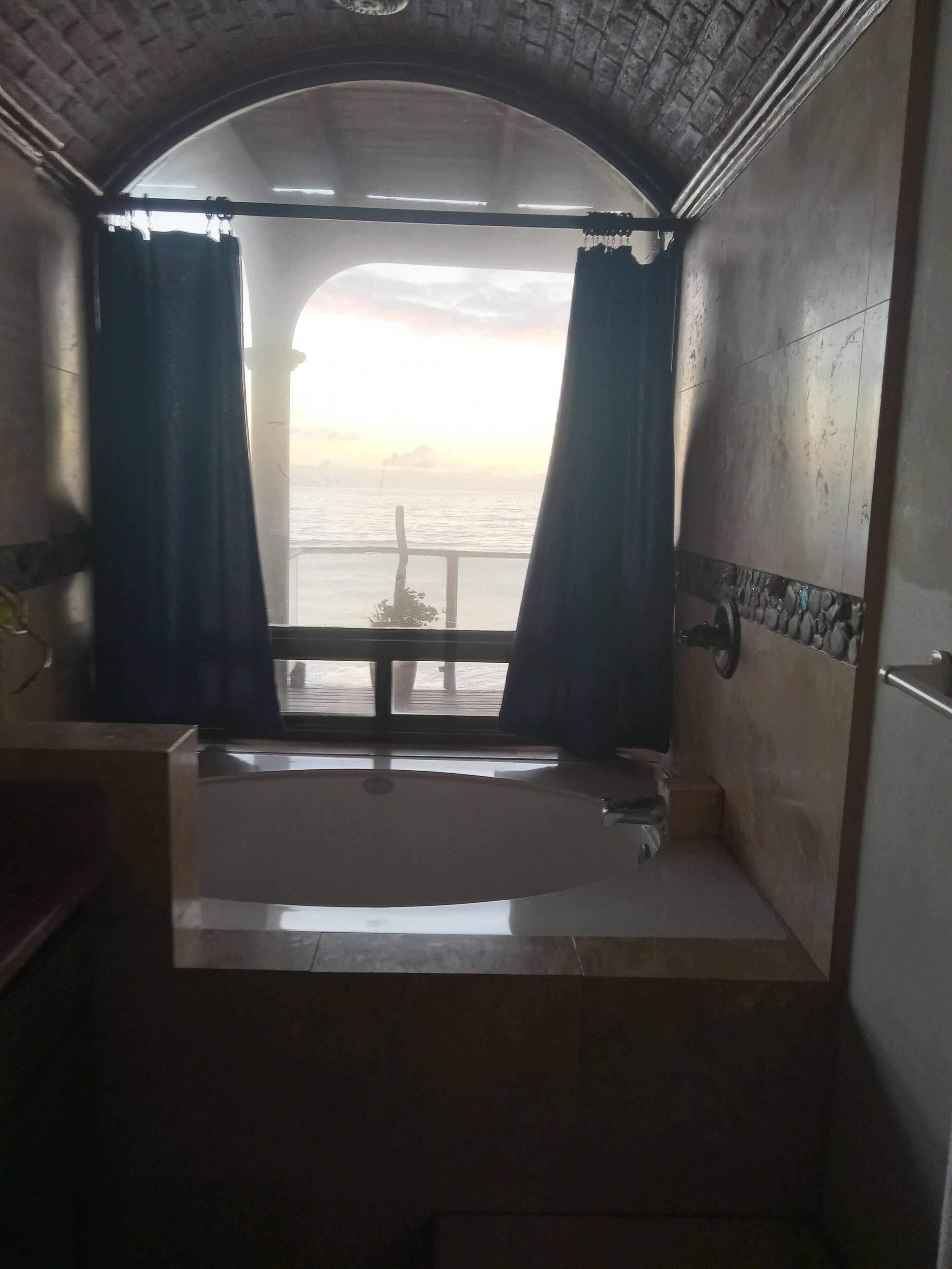 A bathroom with a bathtub, a window with curtains, and a view of the sea.