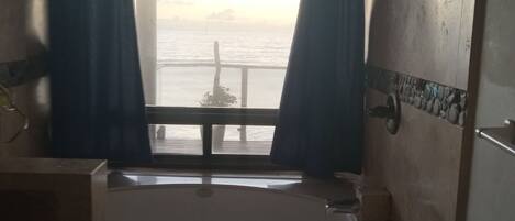 A bathroom with a bathtub, a window with curtains, and a view of the sea.