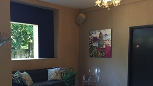 A living room with a dark blue sofa, a glass coffee table, a clear chair, a potted plant, a chandelier, a painting on the wall, and a door. - Palm Residence (Petionville)