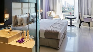 A modern hotel room with a large bed, a glass partition, a sitting area with a table and chairs, and a view of the city. - Eskay Resorts (Mumbai)