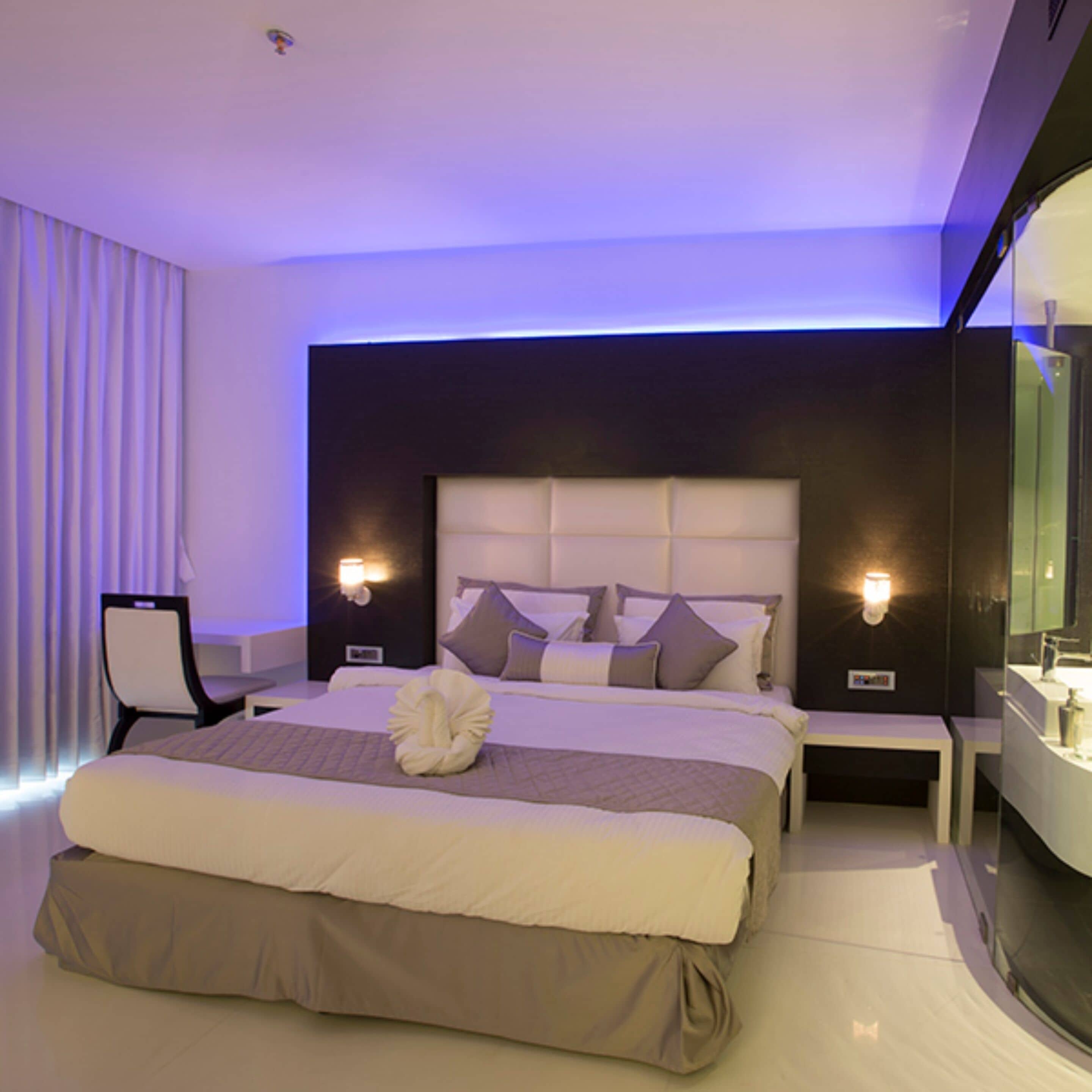 A modern hotel room with a large bed, a bedside table, a chair, and a bathroom with a mirror.