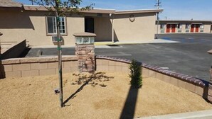 A building with a stone pillar, a small tree, and a paved parking area. - Motel 6 on Maple (Hesperia)