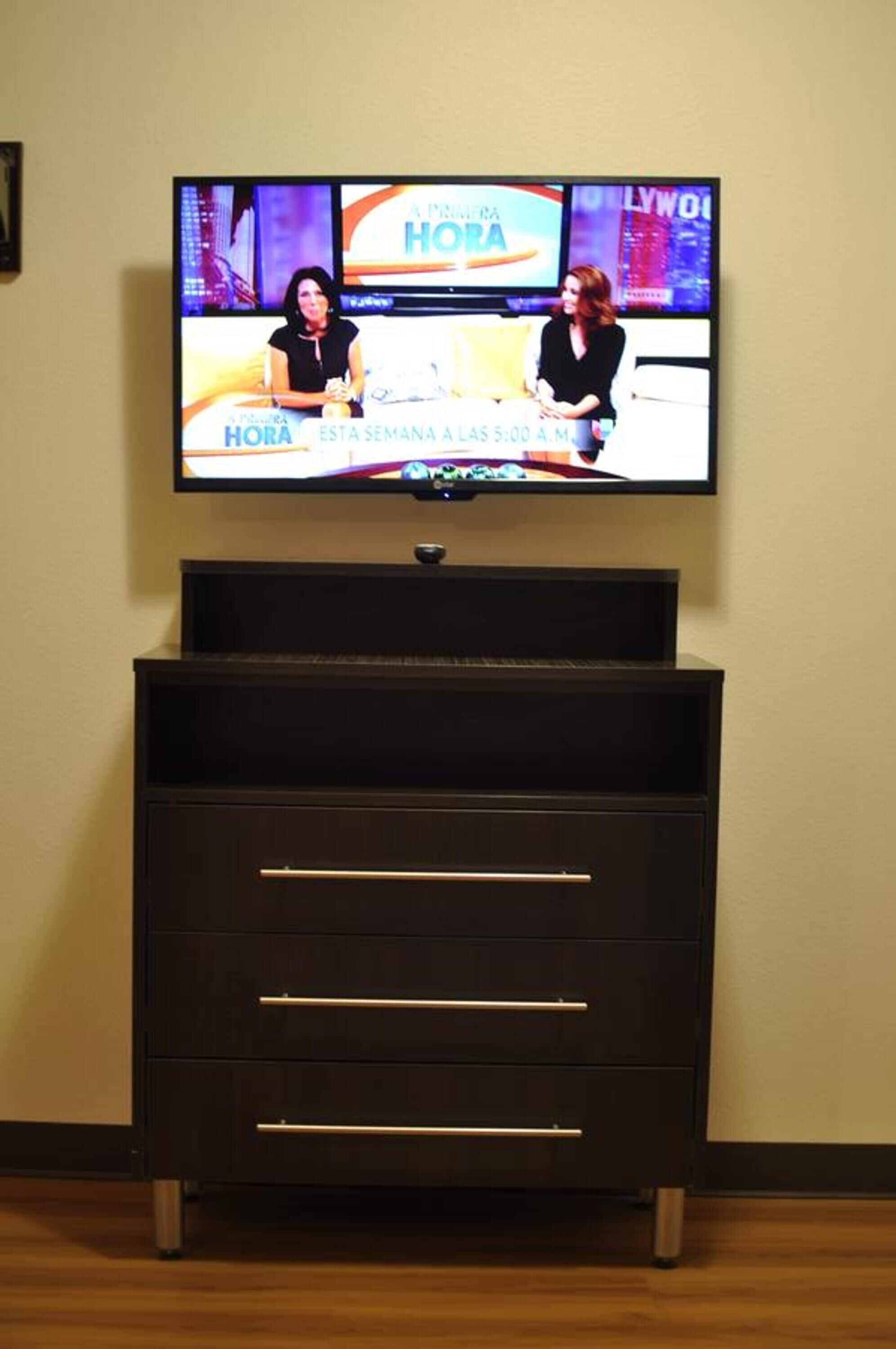 A television mounted on a wall displaying a news program. Below the TV, there is a dark wooden cabinet with drawers.