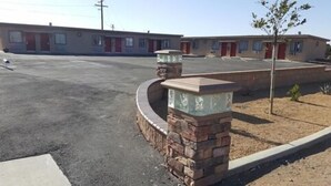 A motel with a parking lot and a small landscaped area with a tree. - Motel 6 on Maple (Hesperia)