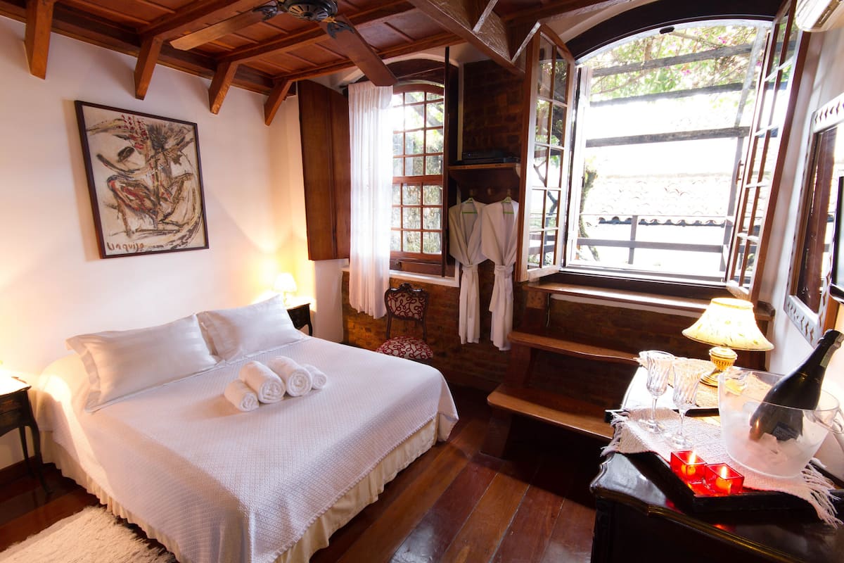 A bedroom with a large bed, a wooden ceiling, a window with curtains, a chair, and a table with a bottle and glasses.