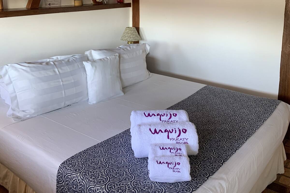 A neatly made bed with white bedding and a patterned throw, a wooden ceiling, and artwork on the walls.