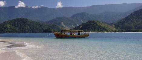 A boat on a clear turquoise sea with a beach and mountains in the background.