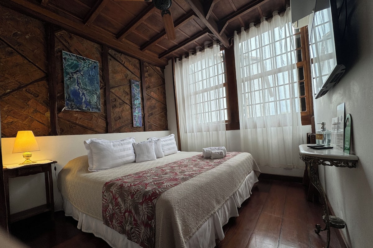 A room with a wooden ceiling, a bed with white bedding, a nightstand with a lamp, and a window with sheer curtains.