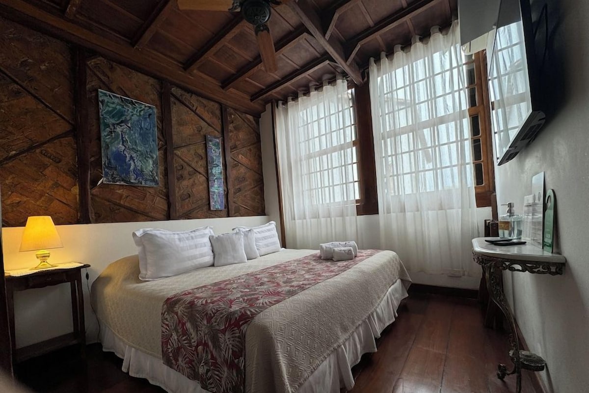 A room with a bed, a nightstand, a lamp, a window with curtains, and a wall clock.