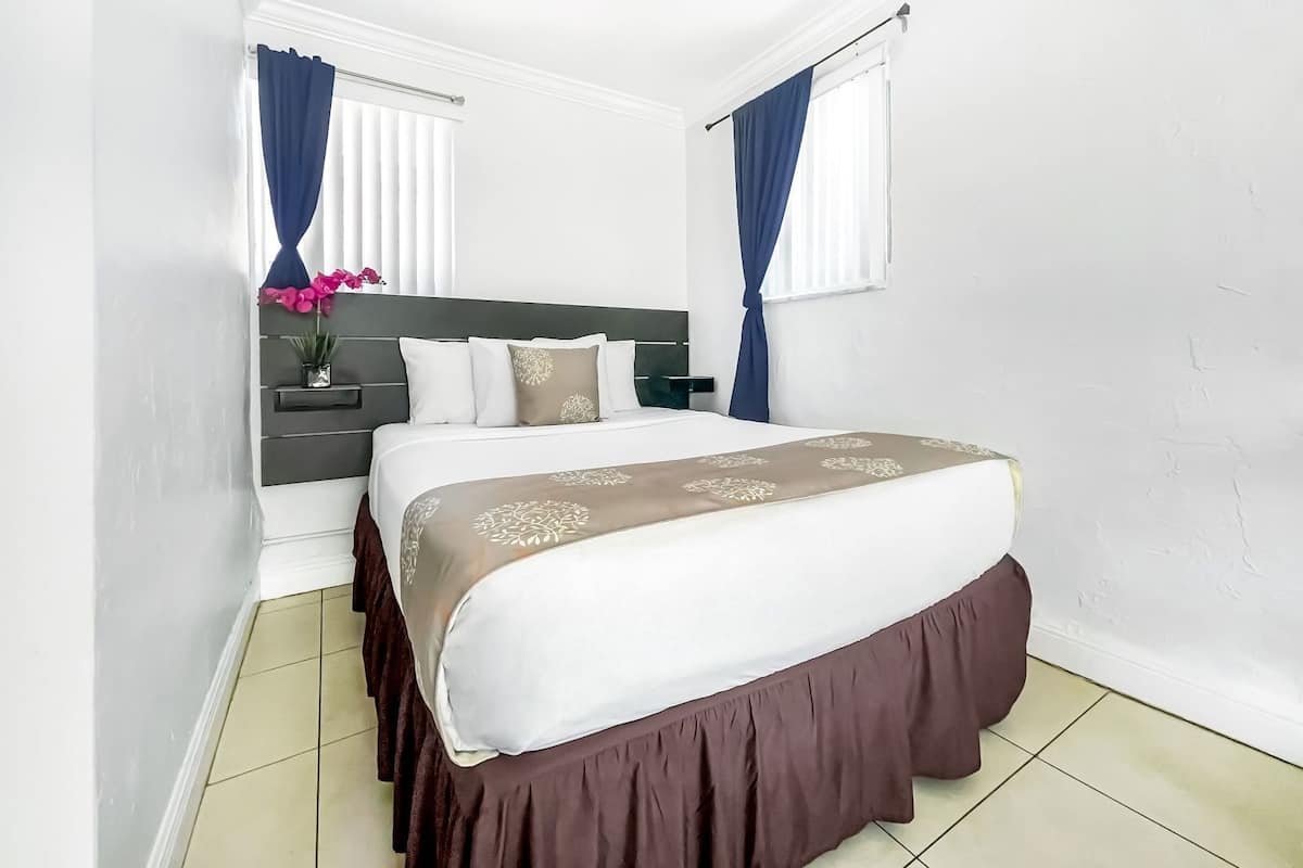 A hotel room with a bed, a bedside table, a nightstand, a window with curtains, and a wall-mounted shelf with a vase of flowers.
